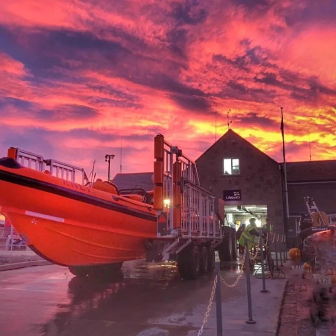 An incredible sunset over the top of one of our RNLI Lifeboat Stations. An inshore lifeboat is outside the station and the sky above is a mix of pink, orange, purple and yellow.
