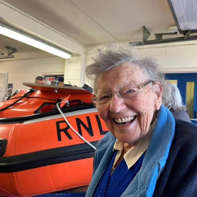 A care home resident enjoying a guided tour around one of our RNLI Lifeboat Stations. She's smiling towards the camera with a lifeboat behind her.

