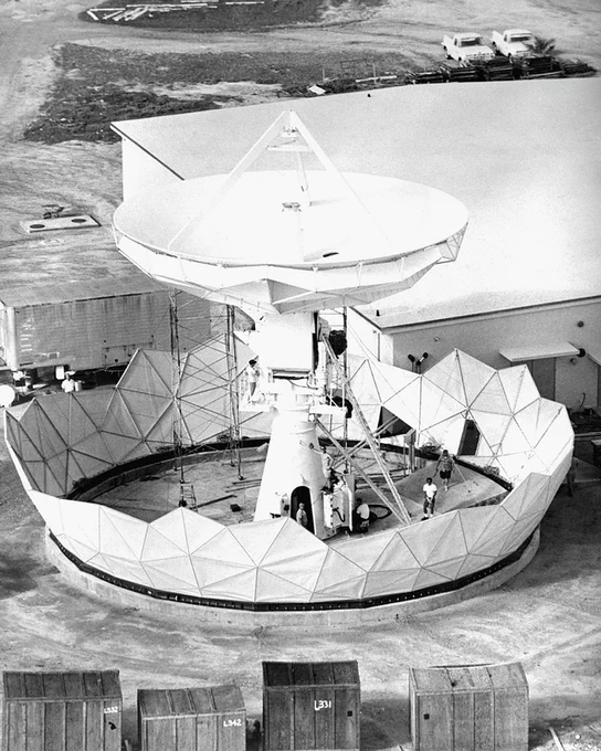 Large satellite dish antenna with a circular base and protective panels, surrounded by several workers performing maintenance or assembly tasks.