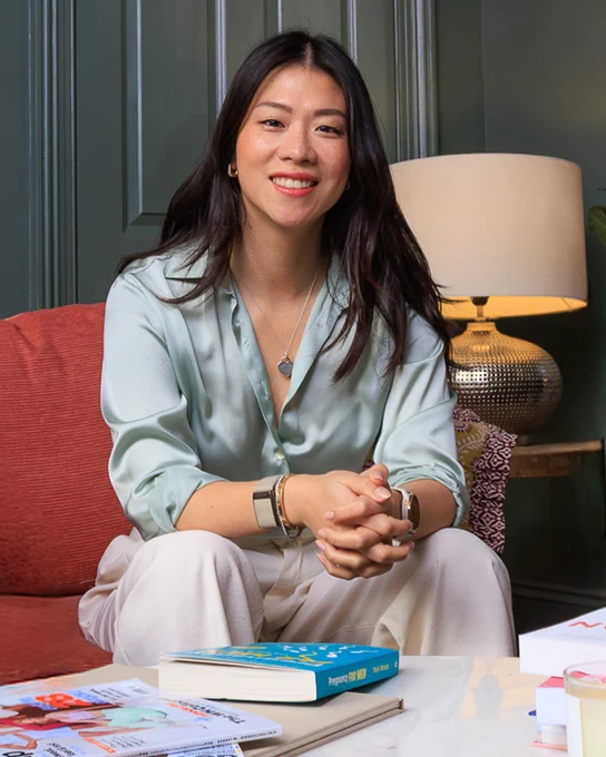 Ambra Zhang,  wearing a silvery grey blouse and pale pink trousers, sits on a red couch with hands clasped. In front are several books on a marble table.