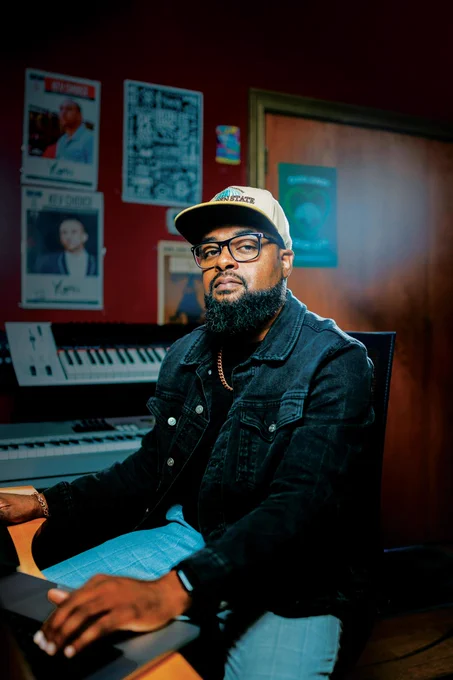 Person sitting at a desk in a music production studio, wearing a denim jacket and a baseball cap, with a keyboard and various awards in the background.