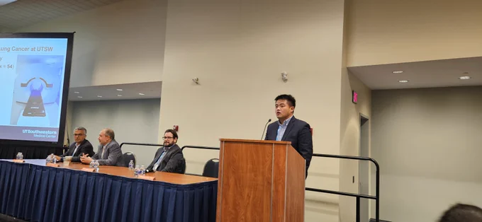 A man in a suit giving a talk behind a podium with a panel of moderators seated next to him.
