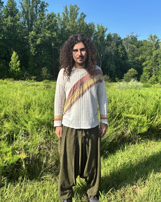 Alej Moses de la Vega, a young man with long, dark curly hair wearing a white shirt with a warm-toned diagonal stripe and green pants, stands in front of a forest.