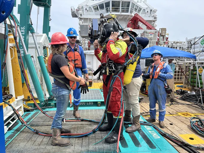 people in hard hats stand around a person in a full-body red and yellow suit with a helmet on the deck of a large research vessel
