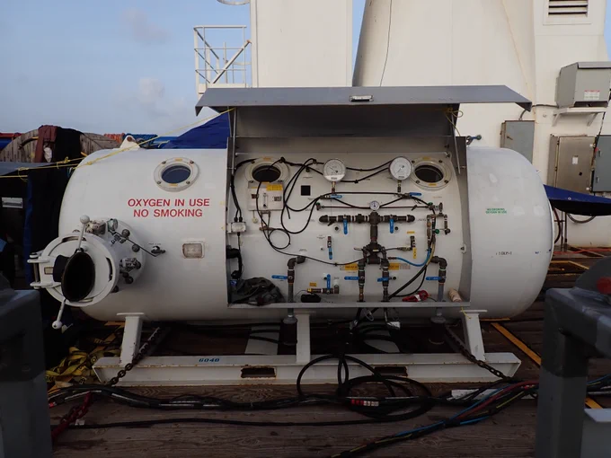 a white, cylindrical object with many pipes, wires, and buttons, and a small port window to view inside the chamber