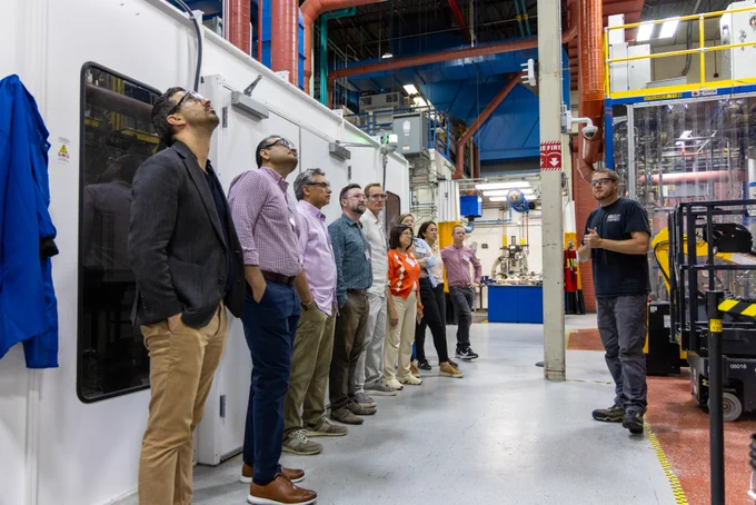A group of eight professionals attentively observing a demonstration inside a lab, with a researcher gesturing toward machinery.