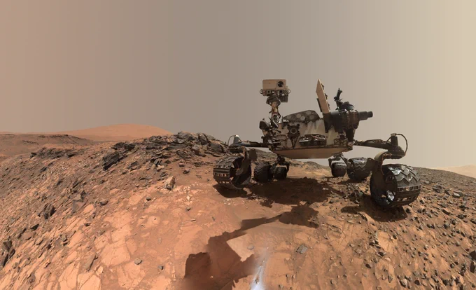 A low-angle self portrait shows the Curiosity rover on a hill on the surface of Mars. The image is color enhanced and gives off a rusty-beige hue. The rover used its MAHLI instrument to take several photos that were then composed into this one shot. The large rover is in the center-right of the image and is slightly dusty. There are dozens of various rocks to the left of it and small hills along the horizon. Inside the right wheel of the rover is a small little rock that tagged along for the ride.