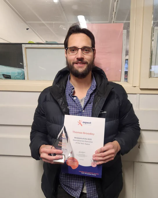 A man smiling while holding a triangular glass award and certificate, standing indoors wearing glasses, a black jacket, and checkered shirt.
