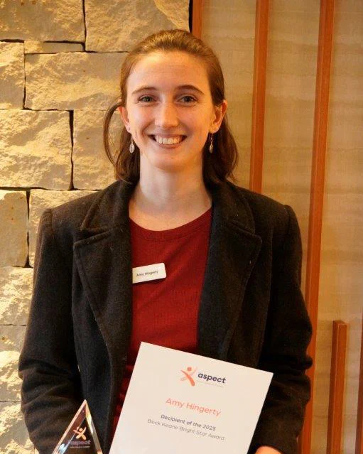 A women smiling while holding a triangular glass award and certificate, standing indoors against a stone wall.