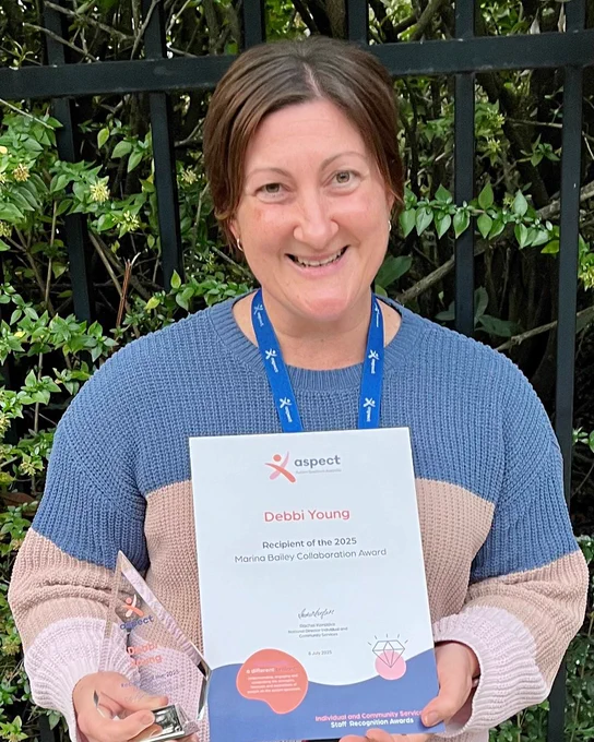 A women smiling while holding a triangular glass award and certificate, standing indoors against a stone wall.