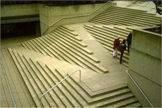 A large concrete staircase with an integrated ramp flanked by shallow, narrow steps that span the width of the structure. Three people are walking up the central ramp, which is embedded within the stair design. The layout is symmetrical but unconventional, blending stairs and ramp in a sculptural, abstract form. Metal handrails are present on the sides.