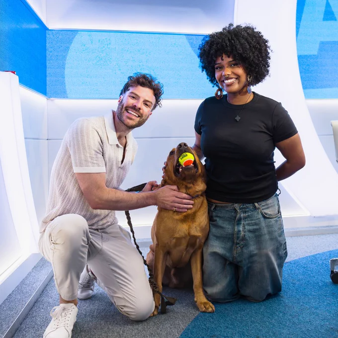 Jordan North kneeling on the floor next to Creed, the Labrador dog, and his owner, Rodas. They're smiling at the camera and Creed has a tennis ball in his mouth. 


