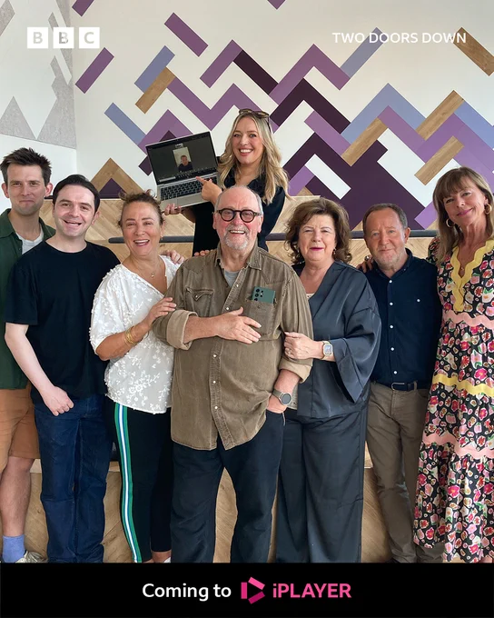 Group photo featuring the cast of the Two Doors Down Christmas Special. One person appears via a laptop. BBC branding frames the image and reads: "Coming to BBC iPlayer."