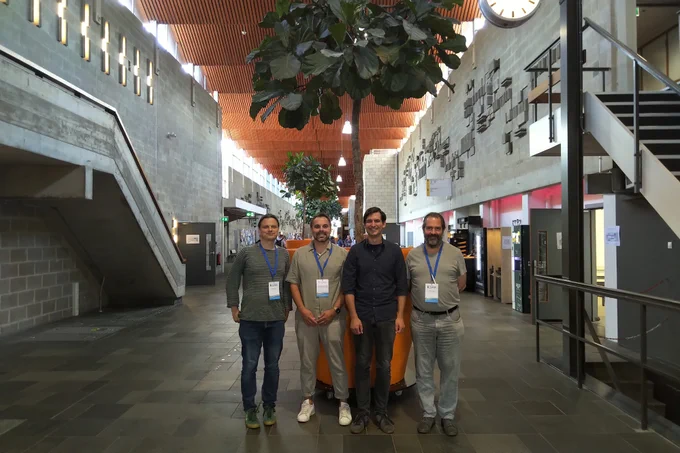 Four ESRA conference participants smiling and standing in a modern hall with concrete walls, a glass roof, large indoor plants, and a clock and staircase in the background. Other attendees are visible further down the hallway.