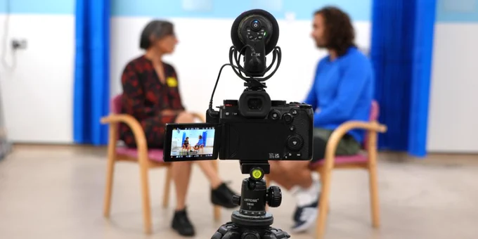 A video camera is in the foreground of the image, with two people in the background, slightly out of focus. They are sitting on chairs and talking to each other. One person is a Trust clincian, the other is celebrity fitness expert Joe Wicks.