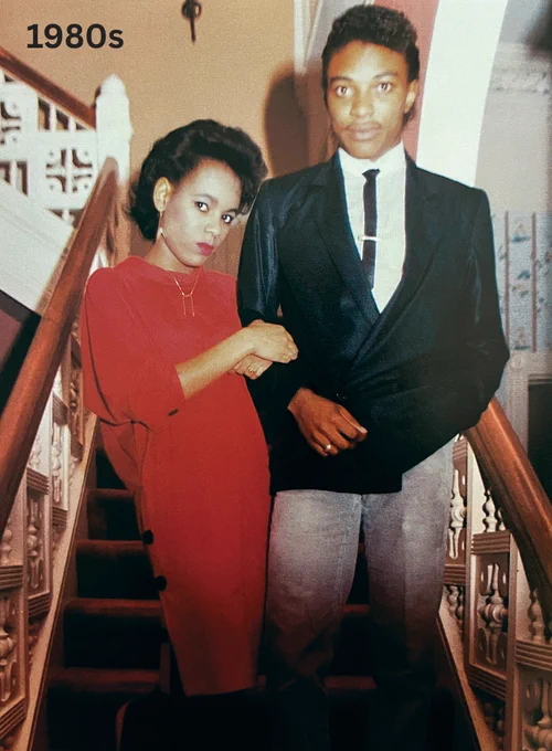 Two people posing on a staircase, the woman in a red dress and the man in a dark blazer over a light shirt and dark trousers, reflecting 1980s fashion.