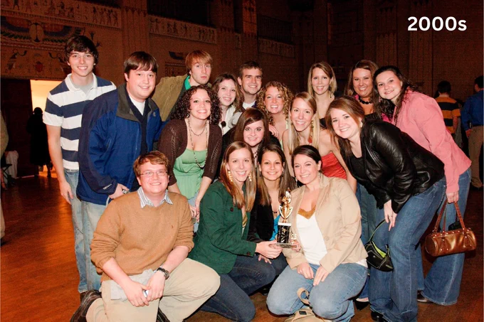 Group of smiling young adults posing in a hall, some holding a trophy, with the text "2000s" on the corner of the photo. 