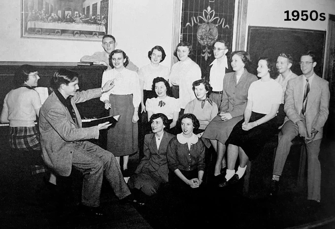 A black and white photograph from the 1950s showing a group of students gathered around a teacher who is seated and holding a book, in a classroom setting.