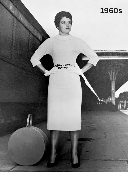 A woman stands on a platform beside a large round suitcase, dressed in an elegant 1960s outfit, featuring a belted waist and a leopard print belt.