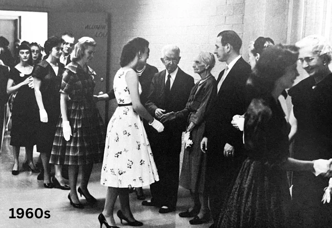 Black and white photo from the 1960s showing a group of college students and administrators in formal attire greeting each other in a hallway.