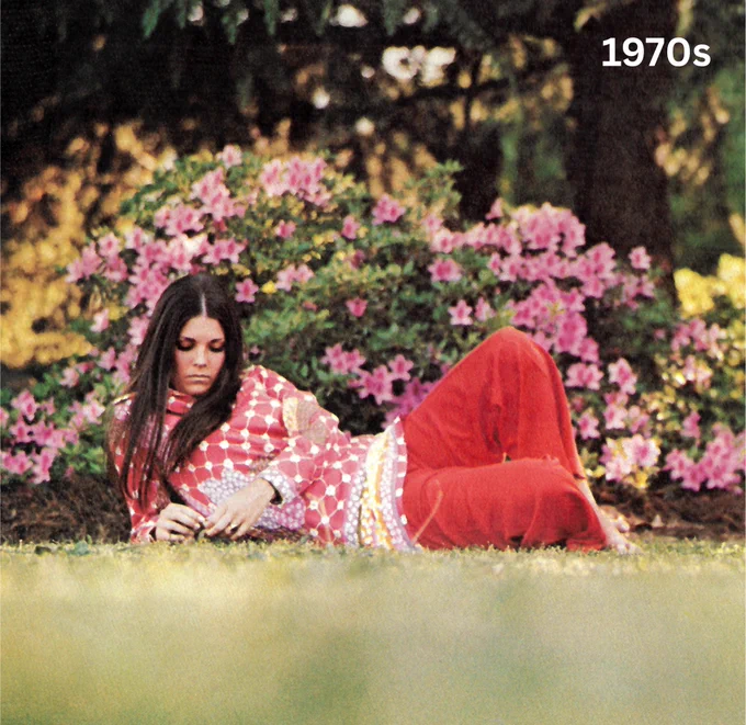 Woman in a vibrant pink patterned outfit and red pants reclining on grass with lush greenery and flowers in the background, evoking a 1970s style.