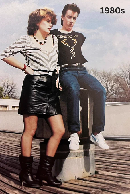Two people dressed in stylish 1980s fashion sit on a ledge. The woman wears a black and white striped top with a black leather skirt and boots, while the the man sports a graphic t-shirt and jeans with white sneakers.