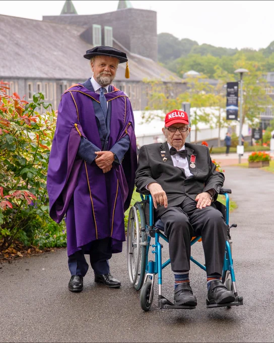 Person in academic regalia standing next to another person in a wheelchair wearing a cap with the word "Nello" outside a building.