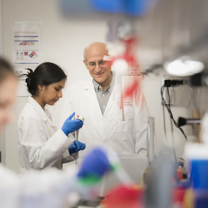 Two researchers in lab coats working in a laboratory, one closely observing a test tube and the other reviewing results. The backdrop features scientific posters and equipment indicative of a scientific research environment.