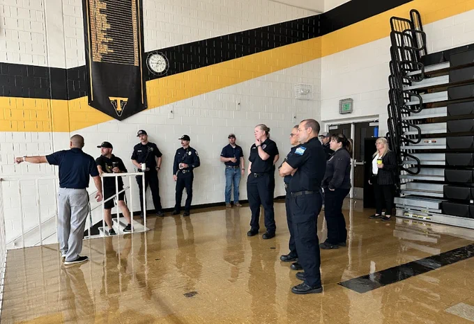 Firefighters and school staff listen during a walkthrough inside a gym with black bleachers and yellow accents.