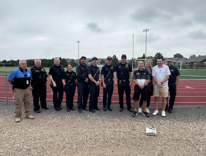 Police, fire, and school staff stand on a track with goalposts and homes in the background under cloudy skies.