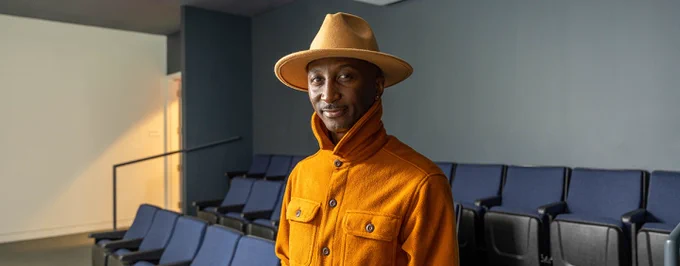 Person in a mustard jacket and matching hat smiling in a room with rows of blue seats.