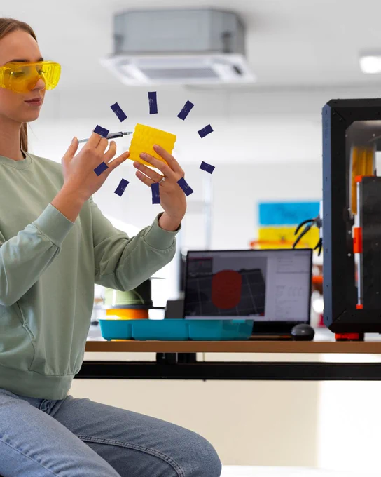 Person examining a yellow 3D-printed object in a lab with a 3D printer and computer visible in the background.
