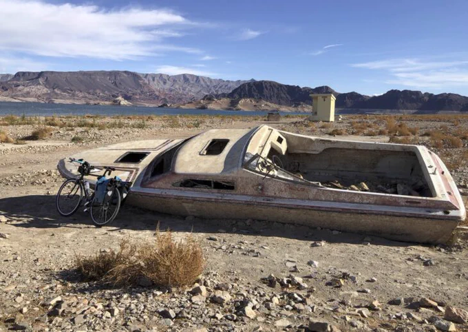 A bicycle leaning against a dirty old wrecked speedboat in front of a desert reservoir.

