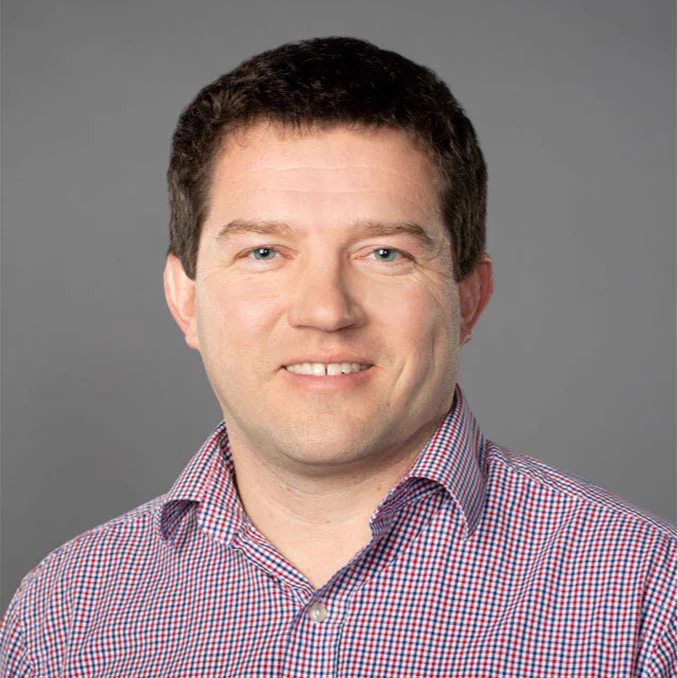 Portrait of a person wearing a checkered shirt, smiling against a gray background.