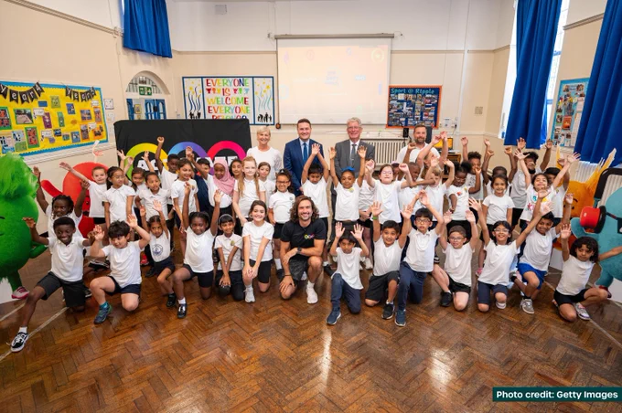 Joe Wicks, Health and Social Care Secretary Wes Streeting, teachers and schoolchildren assembled for a group photograph in a school assembly hall. The children all have their hands in the air and smiles abound.