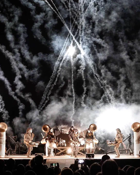 A dynamic live concert scene featuring a stage with a backdrop of dramatic fireworks and smoke effects.