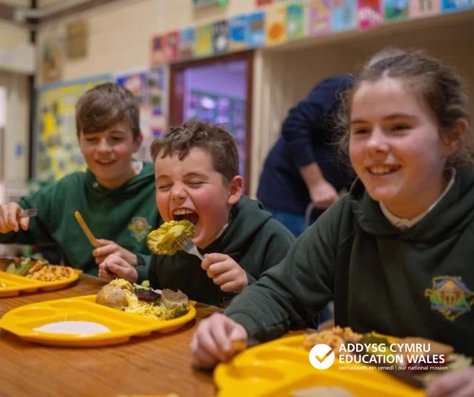 Three children in green school uniforms sitting at a wooden table eating meals from yellow trays in a school cafeteria.