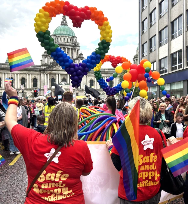 Image is a photo taken at last year’s event. It shows the back of two Action for Children staff wearing red t-shirts which read ‘Safe and happy childhoods’. They are among a crowd of people waving rainbow flags and there is a large heart made out of multicoloured balloons in the sky which frames of the spire of Belfast City Hall.
