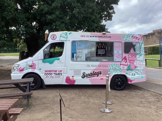 Ice cream van parked under a tree in the school grounds with the Marple Hall School logo on the from of the van.
