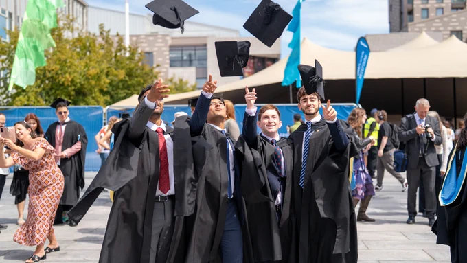 Four university graduates in black gowns joyfully throw their caps into the air on a sunny day. They stand in an outdoor area with a crowd of people in the background. Bright banners and a mix of greenery and urban architecture are visible. 