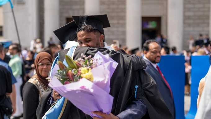 Graduates in black caps and gowns embrace joyfully, one holding a bouquet of colorful flowers. 