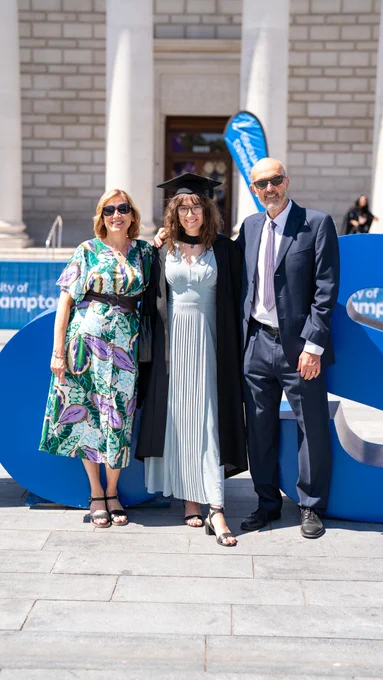 A graduate in academic regalia stands between a woman in a floral dress and a man in a dark suit and sunglasses. They pose in front of large blue letters with a building's stone facade in the background.