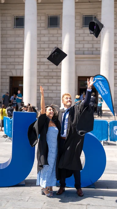 Two graduates celebrate by throwing their caps in the air. They wear black gowns, standing in front of a large blue sculpture. A historic building with columns and a "GRADUATION ENTRANCE" banner is in the background. The day is sunny, with other people around.