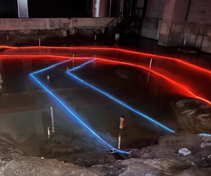 A photograph of the archaeological remains of The Rose Playhouse, currently preserved underwater in a very wide and tall basement underneath an office block, looking down from the modern day viewing platform across the flooded space east to west. Illuminated rope lights in red just underneath the surface of the water outline the position of the inner and outer walls of the seating galleries that ring the yard. Immediately in front of the viewer are two nearly-parallel ropes of blue lights marking the front of the trapezoid-shaped stage in 1587, when the playhouse was first built, and then pushed back a few feet further north (to the viewer's right) when The Rose was expanded in 1592/3. Photo taken 17 July 2025 by Peter Jolly.