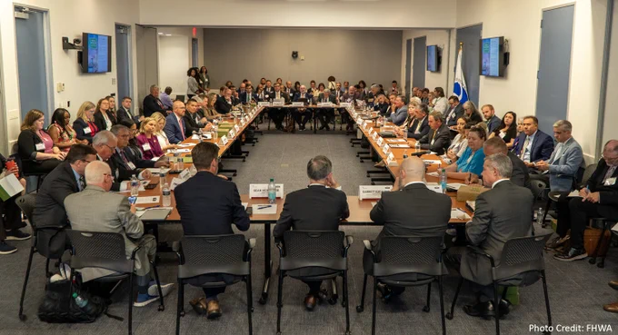 A group of professionals sitting around a large U-shaped conference table during a meeting, with digital screens displaying information on the walls. 