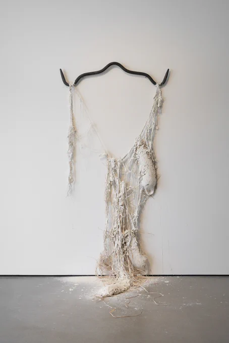 A sculpture hanging against the wall on a metal bar with ships rope and paint hanging down and pooling on the gallery floor.