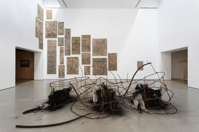 A sculptural installation or rusted metal bars, old rope and charcoal is in the foreground while on the wall behind it sits a series of wooden boxes covered in plant printed material encasing silk worms.