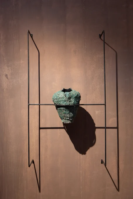 A textured green vessel hangs on a metal frame attached to a pinky-brown wall