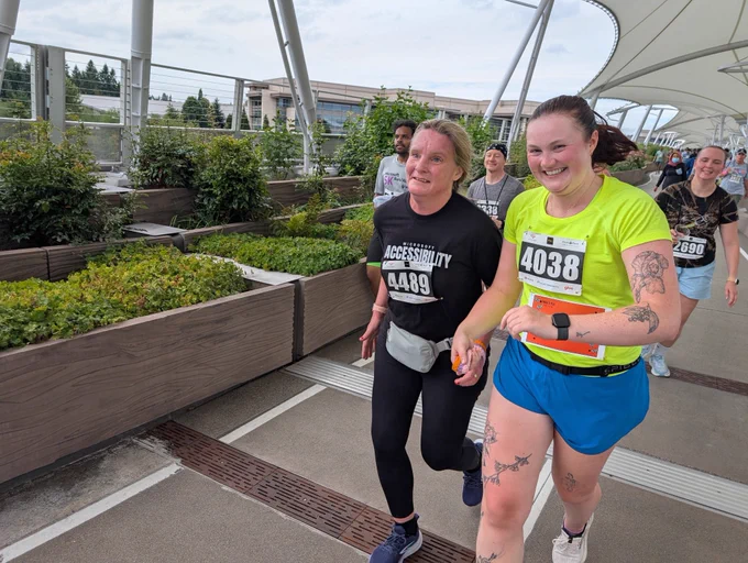 Jessie, who is blind, runs on the Microsoft bridge with her guide Brice. They are connected by a tether, with Brice wearing a bright "GUIDE" shirt. Both are smiling as they participate in the event, surrounded by other runners and greenery.