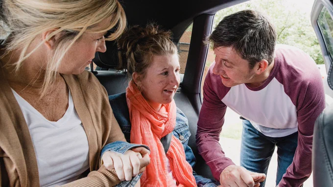 Three people sharing a warm moment in a car, with one person seated while two others lean in supportively from either side.